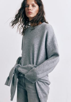Giovane donna con capelli ricci che indossa un maglione grigio oversize annodato in vita e pantaloni grigi, sfondo neutro.