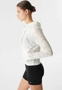 Haut à capuche en maille blanche avec cordon de serrage ajustable, manches longues et poches latérales, associé à un short noir avec un logo sur l'ourlet.