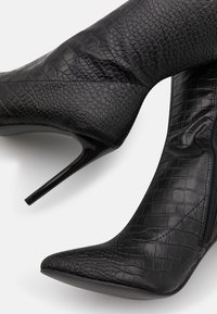 Zapatos de tacón alto de punta negra, hechos de cuero sintético texturizado imitación piel de cocodrilo, que presentan una silueta elegante y altos tacones de aguja.