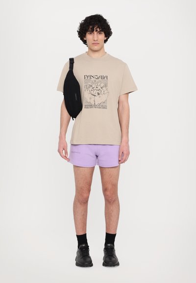 Beige grafisk t-skjorte med en design, sammen med lyse lilla shorts. Svart skulderveske over skulderen, svarte sko og mørke sokker.