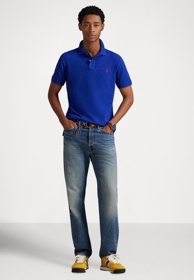 Polo azul royal, jeans azuis desbotados e ténis amarelos. A camisa tem gola, os jeans têm um ligeiro efeito lavado e os ténis têm atacadores.