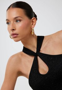 Schwarzes gestricktes Neckholder-Top mit einem Schlüssellochausschnitt, bestehend aus strukturiertem Stoff und einem verknoteten Riemendesign. Goldene Creolen mit Verzierungen.