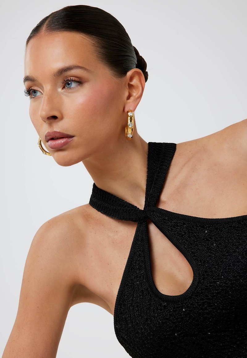 Schwarzes gestricktes Neckholder-Top mit einem Schlüssellochausschnitt, bestehend aus strukturiertem Stoff und einem verknoteten Riemendesign. Goldene Creolen mit Verzierungen.