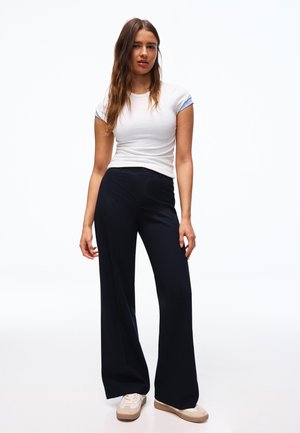 Mlada ženska stoji, oblečena v belo oprijeto majico, široke črne hlače in bež superge, na enobarvnem belem ozadju.