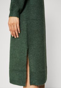 Mano appoggiata su un vestito lavorato a maglia verde scuro con uno spacco laterale di media lunghezza, che mostra parzialmente la gamba.