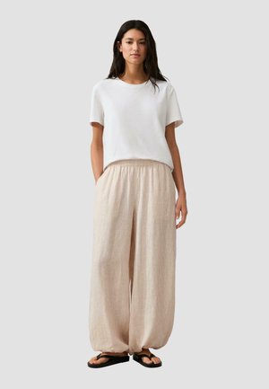 Frau steht und trägt ein weißes kurzärmeliges Hemd, beige Weitbein-Hosen mit elastischen Bündchen und schwarze Flip-Flop-Sandalen, vor einem schlichten Hintergrund.