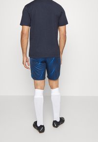 Pantalones cortos atléticos de cuadros azules combinados con una camiseta oscura, calcetas blancas hasta la rodilla y botas de fútbol negras, vistos desde atrás.