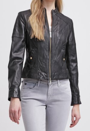 Kvinde iført en tætsiddende sort læderjakke med frontlynlås og lysegrå jeans mod en neutral baggrund.