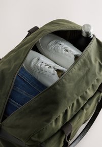 Sac polochon vert olive partiellement dézippé montrant des baskets blanches, un jean bleu plié, et une bouteille d’eau noire avec un bouchon argenté à l’intérieur.