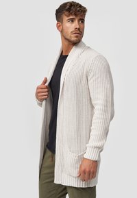 Cardigan in knit crema con apertura frontale, lavorazione a trame e due tasche anteriori. Abbinato a una maglietta nera e pantaloni verde oliva.
