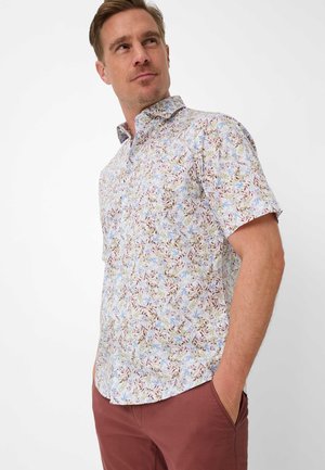 Mann trägt ein kurzärmeliges, durchgeknöpftes Hemd mit einem hellen Blumenmuster und rote Hosen, steht mit einer Hand in der Tasche.