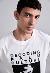 Hvit t-skjorte i bomull med svart grafisk trykk med teksten "DEKODING PALMEKULTUR" og et palmetre-design. Rund hals, korte ermer.