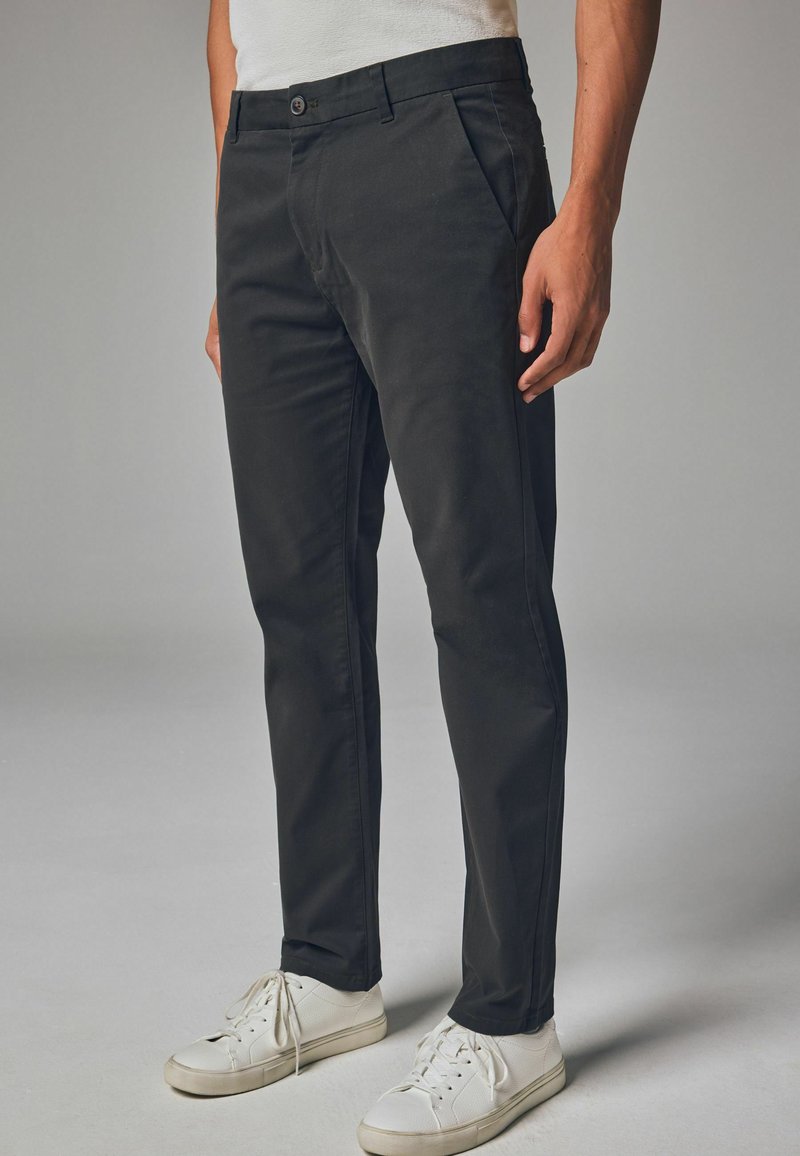 Schwarze Chinos mit geradem Bein, aus einem Baumwollmischgewebe, mit Fronttaschen und einem Knopfverschluss. Weiße Sneakers vervollständigen den Look.