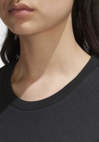 Närbild av en kvinnas nedre ansikte och hals med naturliga hudtoner, iklädd en svart T-shirt med rundad halsringning och med mörkt hår synligt på sidan.