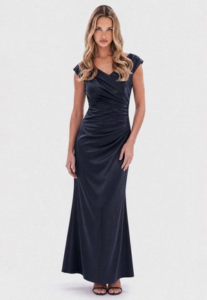 Woman with long wavy blonde hair wearing a sleeveless dark navy satin floor-length gown with ruched detailing and black open-toe heels.