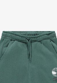 Groene joggingbroek met een elastische tailleband en trekkoord, zijzakken en een wereldbolafbeelding aan de zijkant. Zacht, textuurstof.