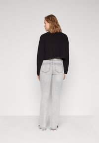 Svart, kort strikkegenser, lys grå utsvingte jeans med baklommer, og svart-hvite mønstrede joggesko. Modellen er sett fra bak.