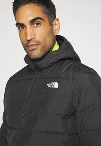 Svart puffjacka med huva, med quiltat mönster och en vit logotyp på bröstet. Bärs över en klar grön topp.