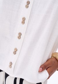 Losse witte blouse gemaakt van textiel met een structuur, met houten knopen aan de voorkant en een zoom die zachtjes omhoog loopt.