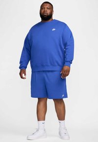 Felpa blu Nike con maniche lunghe e collo a giro, abbinata a pantaloni corti blu coordinati. Logo Nike bianco presente su entrambi i capi.
