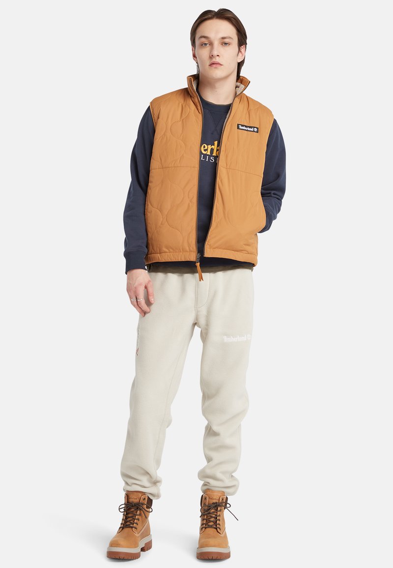 Gilet marrone imbottito sopra una felpa blu scuro, abbinato a pantaloni in pile chiari e scarponi da trekking color sabbia. Logo ben visibile sul gilet.