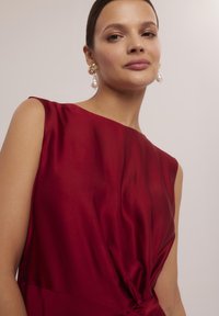 Czerwona satynowa sukienka o bezrękawowym fasonie i szczególe w postaci zawiązania w talii. Modelka uzupełniła stylizację złotymi kolczykami z perłami. Gładka tekstura.