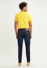 Hombre con cabello rizado que lleva una camiseta amarilla, jeans azul oscuro y zapatillas amarillas, de pie de espaldas sobre un fondo liso.