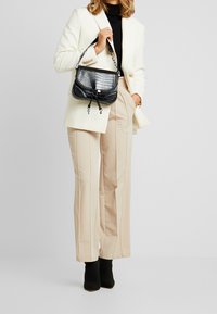 Svart handväska med krokodilprägling, kedjetillbehör och dragskoöppning, bärs med en outfit bestående av en vit blazer och beige byxor.