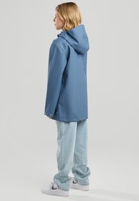 Junge Person mit schulterlangem blondem Haar, die einen blauen Kapuzenregenmantel, hellblaue Jeans und weiße Turnschuhe trägt und seitlich steht.