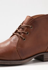 Stivale alla caviglia in pelle marrone con punta rotonda, tre occhielli per allacciatura frontale e tacco basso impilato su sfondo bianco.