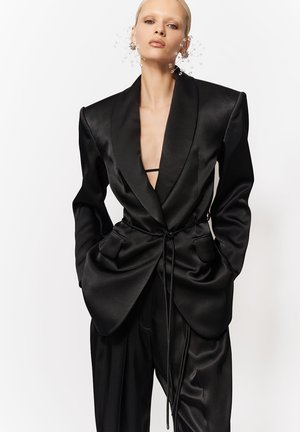 Femeie purtând un sacou supradimensionat din satin negru, cu reveruri late, cordon strâns în talie, pantaloni asortați și cercei mari decorativi cu motive florale.