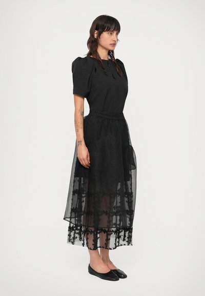 Melna kleita ar pieguļošu augšu, īsām puff piedurknēm un caurspīdīgu, izšuvumu tilla svārku. Akcentēta ar dekoratīviem elementiem gar apakšmalu.