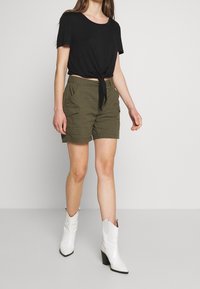 T-shirt noir court avec un nœud à l'avant, short cargo vert olive avec poches latérales, et bottes de cowboy blanches à talons.