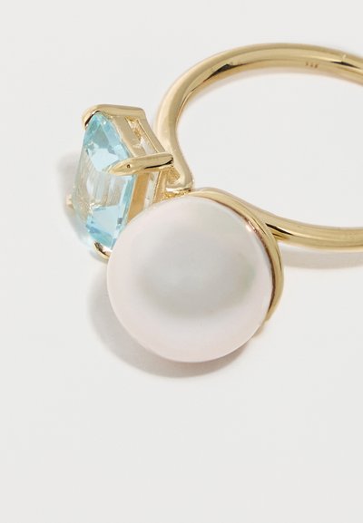 Bague en or ornée d'une grande perle blanche ronde et d'une pierre précieuse de coupe carrée bleu clair, placées côte à côte sur un fond blanc uni.