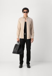 Camicia beige con bottoni, canotta nera, jeans neri, stivaletti neri, che porta una borsa a tracolla nera con texture, indossando occhiali da sole scuri.