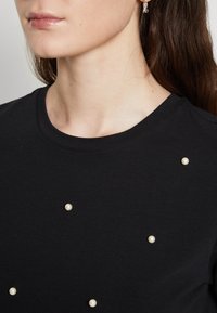 Camiseta negra de tejido suave, con escote redondo y cuentas blancas decorativas colocadas aleatoriamente en el frente.