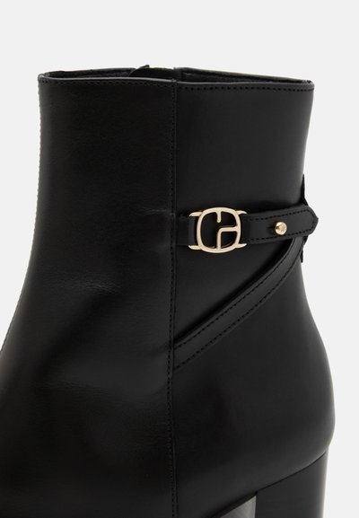 Bottine en cuir noir avec une boucle dorée décorative et un détail de sangle, présentant un haut talon bloc sur fond uni.