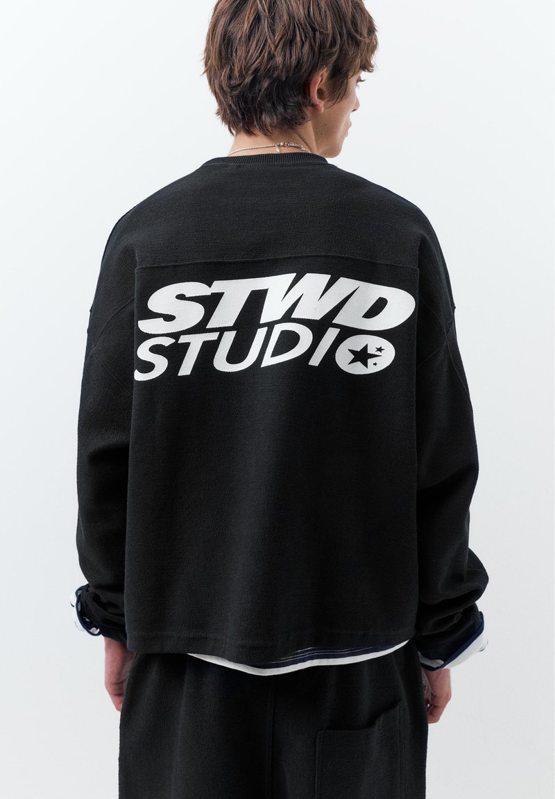 Personne portant un haut à manches longues noir avec le texte blanc audacieux "STWD STUDIO" et un graphique d'étoile au dos, debout devant un fond uni.