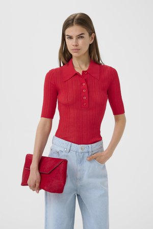 Kobieta w czerwonej prążkowanej koszulce polo z krótkim rękawem, jasnoniebieskich dżinsach z wysokim stanem, trzymająca czerwoną kopertówkę, stoi na tle jednolitego tła.