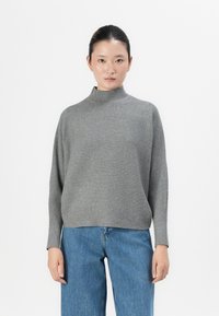 Woman with dark hair tied back, wearing a ribbed grey turtleneck sweater and blue jeans, standing against a plain white background.