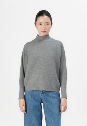 Kvinne med mørkt hår samlet, iført en ribbet grå høyhalset genser og blå jeans, stående mot en ensfarget hvit bakgrunn.
