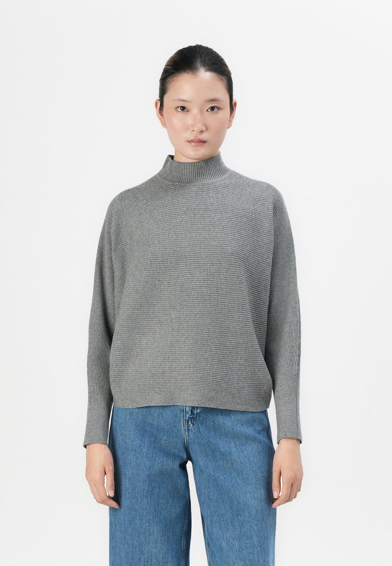 Woman with dark hair tied back, wearing a ribbed grey turtleneck sweater and blue jeans, standing against a plain white background.