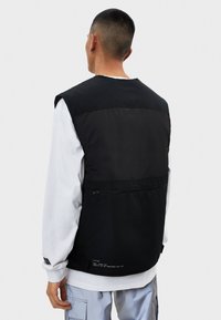 Chaleco negro sin mangas con textura suave, con un bolsillo con cremallera en la parte posterior y una camisa interior blanca en contraste debajo.
