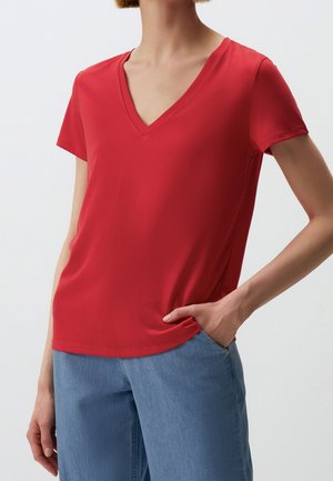 Vrouw draagt een rood shirt met korte mouwen en V-hals en lichtblauwe spijkerbroek, met één hand in de spijkerbroekszak tegen een effen achtergrond.