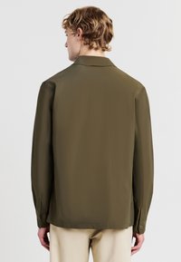 Camicia verde oliva a maniche lunghe con taglio rilassato, colletto a punte e polsini con bottone. Texture liscia, design minimale e senza motivi visibili.