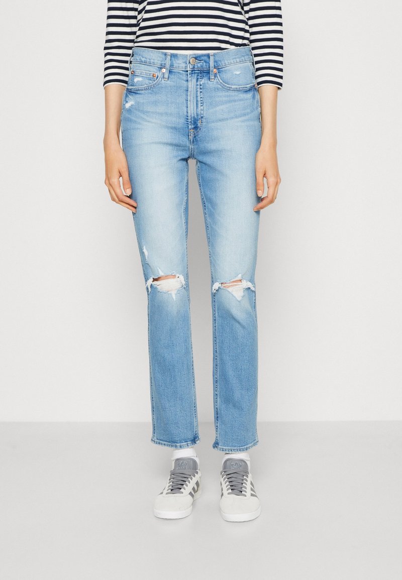 Personne portant un jean bleu clair déchiré, un t-shirt à manches longues rayé, des chaussettes blanches et des baskets blanches et grises se tenant sur un fond uni.