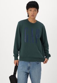 Sweatshirt verde cu guler rotund și logo-ul bleumarin "GAP", din material de bumbac, cu manșete și tiv cu nervuri, combinat cu blugi de culoare albastru deschis și o geantă neagră.