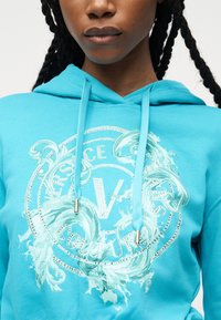 Γυναίκα με πλεγμένα μαλλιά, ντυμένη με φωτεινή μπλε φούτερ με κουκούλα, διακοσμημένη με γραφικό σχήμα νερού σε στροβιλισμούς και με το κείμενο «Versace Couture» ανάγλυφο στο μπροστινό μέρος.