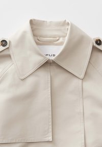 Trench coat beige con un ampio colletto, bottoni doppi e chiusura con zip. Realizzato in tessuto liscio con dettagli di cucitura visibili.