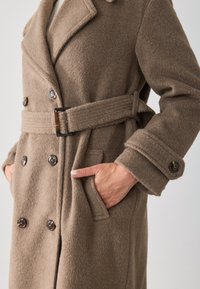 Dubbelzijdige bruine wollen jas met grote knopen, een ceintuur in de taille en zijzakken met omslagen voor extra detail.